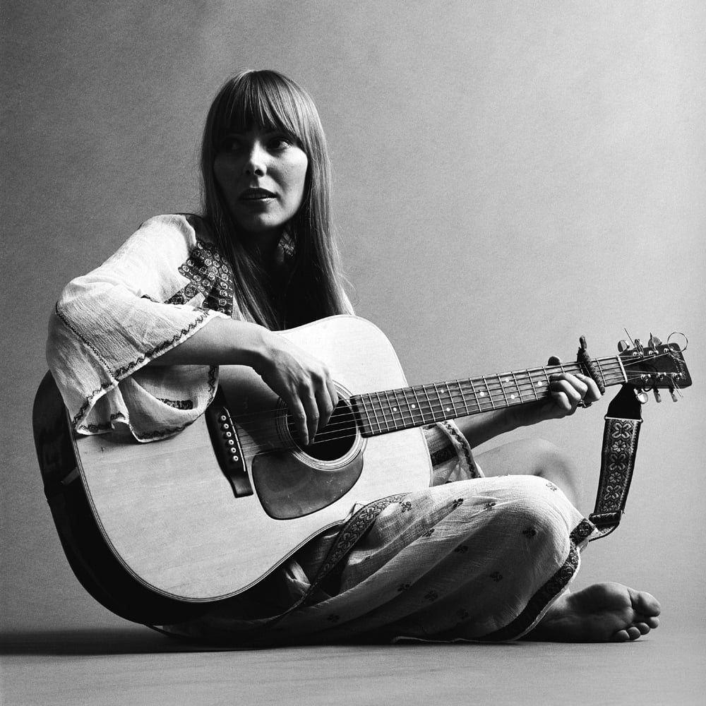 Portrait de la musicienne canadienne Joni Mitchell jouant de la guitare acoustique, en novembre 1968. Cette image est tirée d'une séance de photos pour le magazine de mode Vogue. Mitchell porte une robe blanche ample © Photo par Jack Robinson/Getty Images. 
