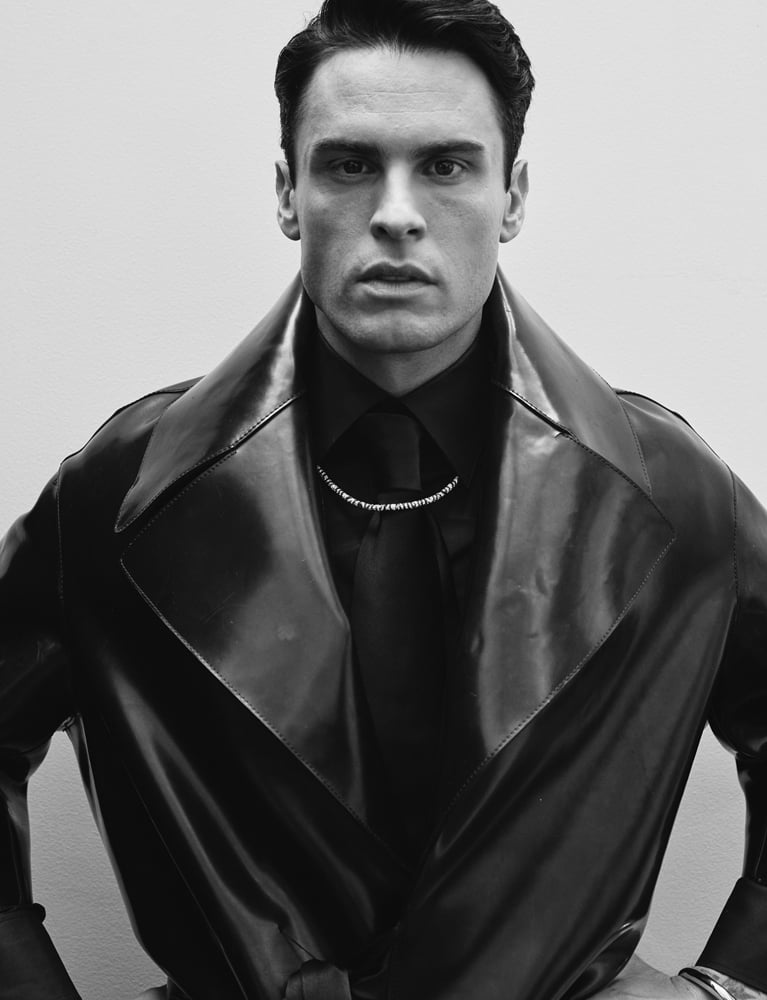 Baptiste Giabiconi en manteau croisé en caoutchouc, chemise en popeline de coton et cravate Saint Laurent par Anthony Vaccarello, collier “Zèbre” et bracelet “Jonc” APM Monaco.