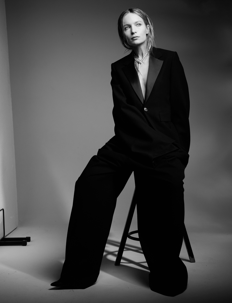 Cliché de l'actrice Diane Rouxel en noir et blanc, vêtue d'un costume masculin noir assise sur un tabouret.