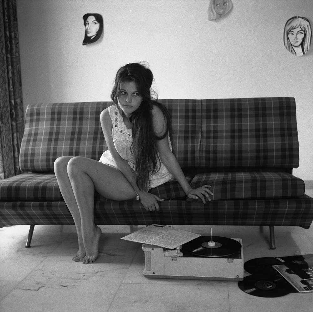 L'actrice italienne Claudia Cardinale assise sur un canapé à carreaux, écoutant des vinyles d'Ella Fitzgerald, avec des visages dessinés accrochés au mur du fond, Rome (1959)