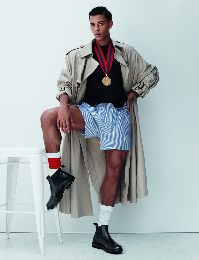 portrait du danseur étoile Guillaume Diop porte un trench en soie de coton Saint Laurent par Anthony Vaccarello, un tee-shirt en jersey de coton et caleçon en seersucker Uniqlo, une médaille Balenciaga, des chaussettes Falke et des boots Church's.