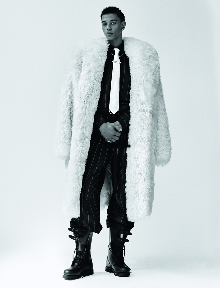 portrait du danseur étoile Guillaume Diop porte un manteau croisé en peau lainée, une chemise et pantalon en flanelle et une ceinture Dolce & Gabbana. Des boucles d'oreilles et bagues Cartier. Une cravate Cinabre, une pince à cravate Dary's et des boots vintage.